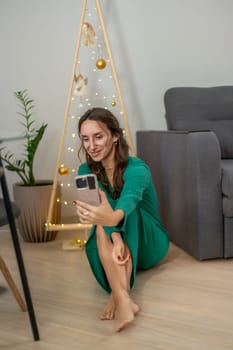 Woman in green dress sits barefoot on floor, smiling at phone, beside a minimalist gold Christmas tree.