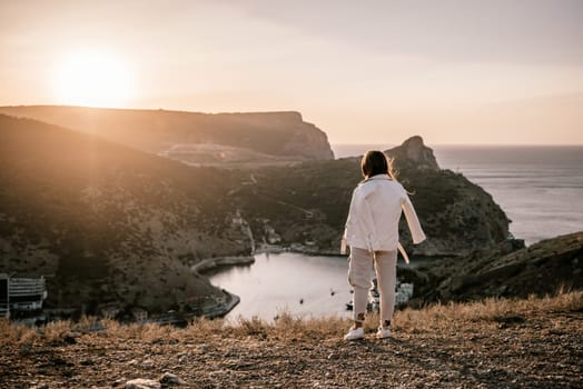 A woman stands on a hill overlooking a body of water. The sun is setting in the background, casting a warm glow over the scene. The woman is enjoying the view and taking in the beauty of the landscape