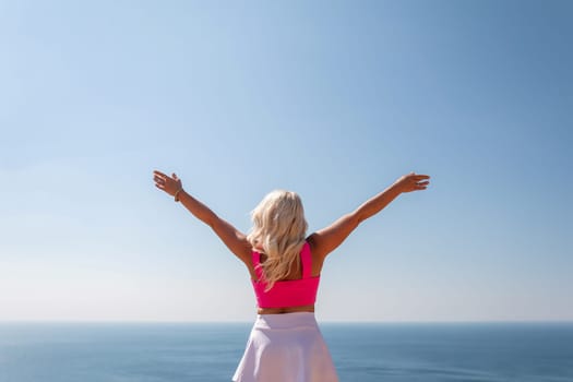 A woman in a pink top and white skirt is standing on a cliff overlooking the ocean. She is smiling and she is enjoying the view