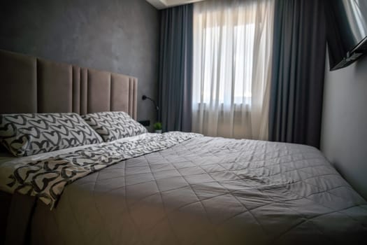 Modern bedroom with a double bed, patterned bedding, textured headboard, dark walls, sheer curtains, and a glimpse of a television.