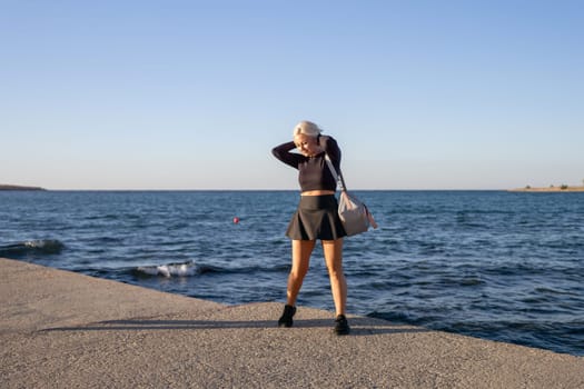 A blonde woman in a black crop top and skirt stands on a concrete pier overlooking a blue ocean. She has a bag slung over her shoulder and is holding her hair. The sky is blue and there are waves crashing on the shore.