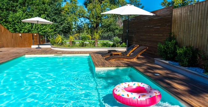 Modern backyard swimming pool with turquoise water, wooden deck, umbrellas, lounge chairs, and pink donut inflatable