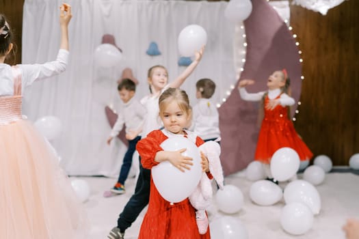 Little girl with a white balloon in her hands walks across the stage past children playing. High quality photo