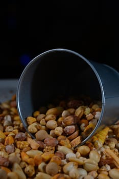 A silver bucket lies on its side, pouring out a medley of nuts, seeds, and colorful snacks onto a dark surface. The mixture showcases an inviting array of earthy tones and textures.