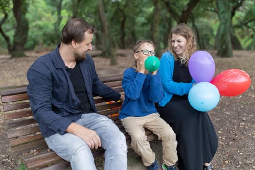 Smiling family having fun in park, child inflating colorful balloon with parents on bench