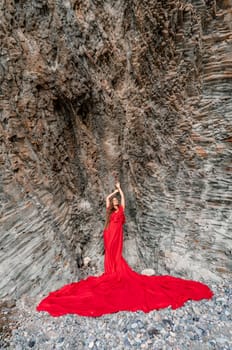 woman sea red dress. Woman with long hair on a sunny seashore in a red flowing dress, back view, silk fabric waving in the wind. Against the backdrop of the blue sky and mountains on the seashore
