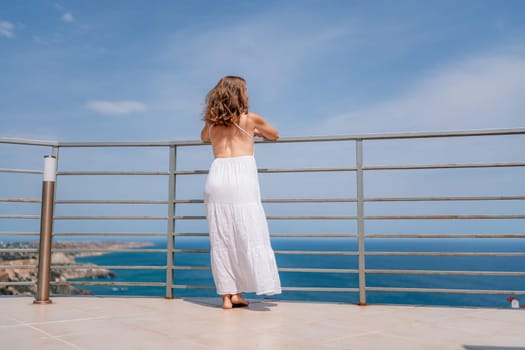 A woman in a white dress overlooks the blue ocean from a balcony with metal railings on a sunny day. The view stretches across the water.
