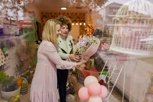 Inside a charming flower shop, two women share smiles as one presents a beautifully arranged bouquet, surrounded by colorful decor and seasonal touches in the background.