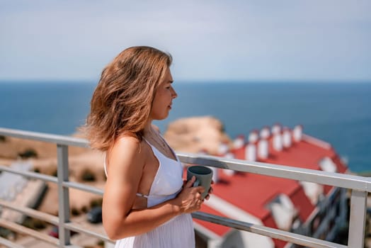 Woman in white dress on balcony, holding a mug, gazes towards the ocean. Red-roofed buildings in background under a sunny sky.