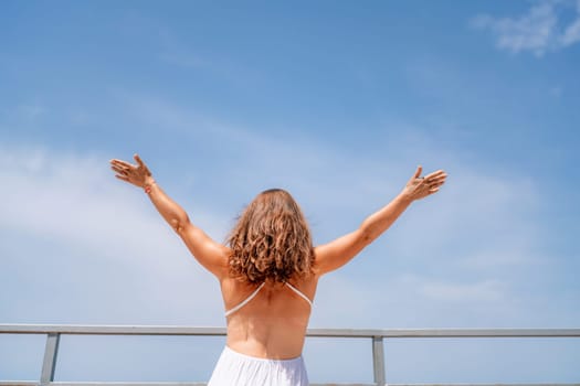 Woman facing away, arms outstretched against a bright blue sky. White dress, curly hair, and distant railing visible. Sunny day.