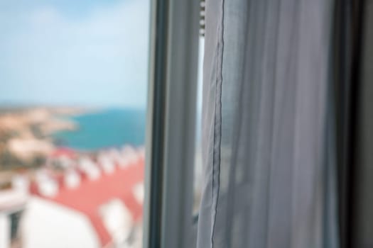 View from a window: A blurred landscape of buildings and blue water seen through a gap in white curtains.