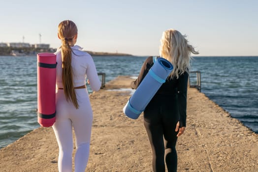 Two women in athletic wear walk along a pier, carrying yoga mats, towards the ocean.