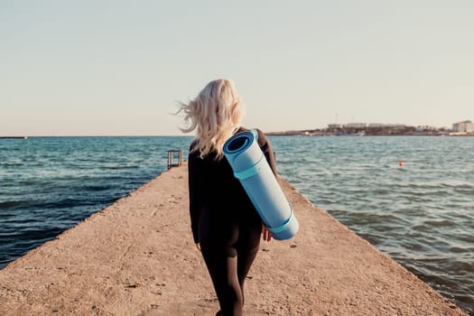 A woman with long blonde hair walks towards the ocean with a yoga mat slung over her shoulder. The sun is shining and the water is calm.