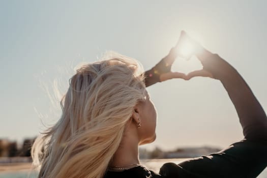 A woman with long blonde hair looks up at the sun, her hands framing the bright light in a heart shape. The sun shines through her fingers, casting a warm glow on her face.