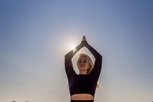 A woman with blonde hair practices yoga outdoors, her hands raised above her head in a prayer position, with the sun shining brightly in the background.