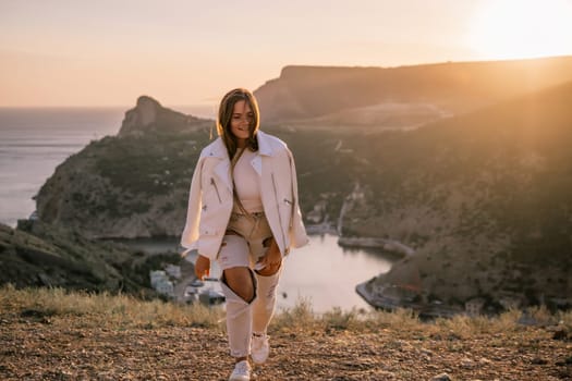 A woman stands on a hill overlooking a body of water. The sun is setting in the background, casting a warm glow over the scene. The woman is enjoying the view and taking in the beauty of the landscape