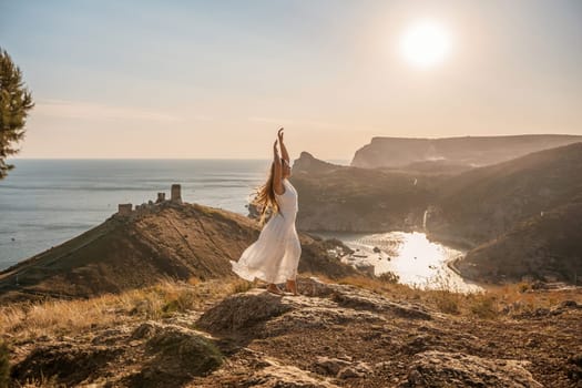 woman stands on a rocky hill overlooking a body of water. She is wearing a white dress and she is in a state of joy or celebration. Concept of freedom and happiness