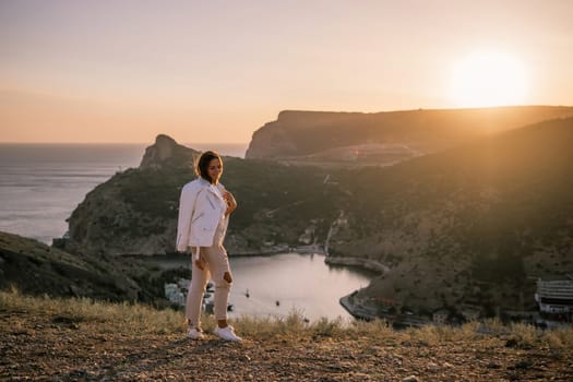 A woman stands on a hill overlooking a body of water. The sun is setting in the background, casting a warm glow over the scene. The woman is enjoying the view and taking in the beauty of the landscape