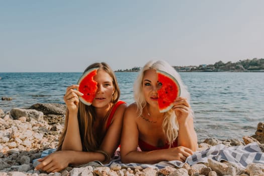 Two women lie on a rocky beach, partially obscured by watermelon slices held to their faces. Bright sun, clear blue sea in the background. Summer fun.