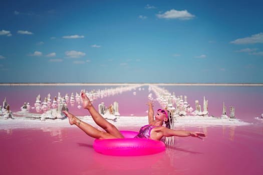 Woman in pink bikini floats on pink inflatable ring in pink lake, surrounded by salt formations. Bright sun, clear sky.