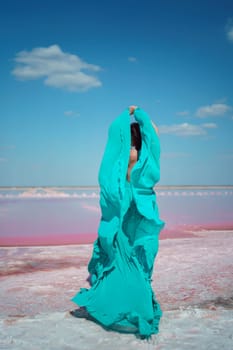 Woman in teal fabric, arms raised, stands on pink salt lake shore under a vibrant blue sky. Windblown fabric billows dramatically.