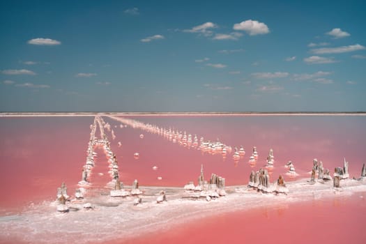 Aerial view of a pink salt lake. White salt formations, resembling crystalline structures, protrude from the pink water. A clear sky with scattered clouds is visible above.