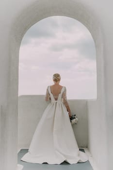 A bride stands in front of an archway, holding a bouquet of flowers. The archway is white and the sky is cloudy. The bride is wearing a white dress and her hair is styled in a bun