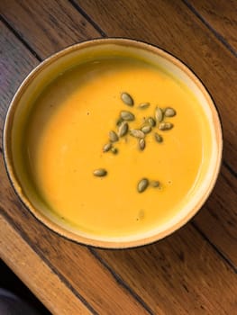 Creamy pumpkin soup served in a bowl, garnished with pumpkin seeds on top. The soup is placed on a wooden table. Photo taken with a smartphone.