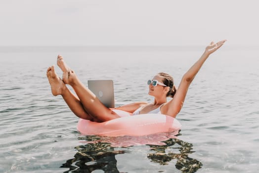 A woman is floating on a pink inflatable raft in the ocean while holding a laptop. Concept of relaxation and leisure, as the woman is enjoying her time in the water while working on her laptop
