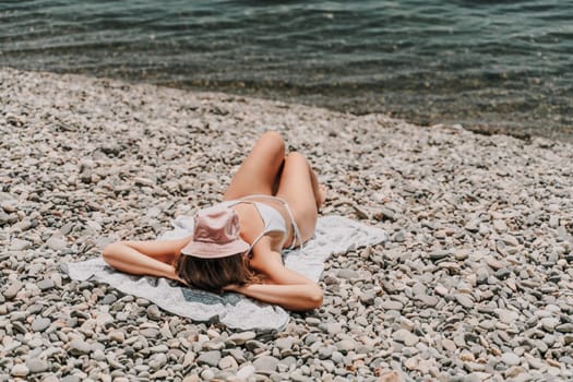 A woman is laying on the beach with a pink hat on her head. She is wearing a white bikini and is laying on a towel. The beach is rocky and the water is calm. Scene is relaxed and peaceful