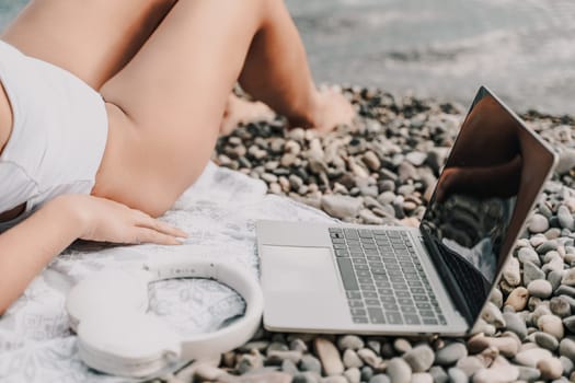 A woman is laying on the beach with a laptop and headphones. The laptop is open and the headphones are on her head. Concept of relaxation and leisure