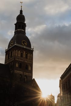 Historic Church Tower with Sunset in the Background.A striking view of a historic church tower silhouetted against a dramatic sky at sunset. The golden sunlight peeks through surrounding buildings, casting a warm glow