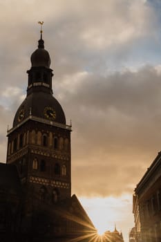 Historic Church Tower with Sunset in the Background.A striking view of a historic church tower silhouetted against a dramatic sky at sunset. The golden sunlight peeks through surrounding buildings, casting a warm glow