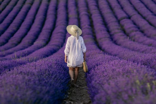 A woman is walking through a field of purple flowers. She is wearing a white shirt and a straw hat