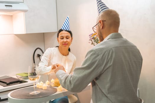 Here's a caption for the image: A man and woman, both wearing party hats, celebrate a birthday. They're in a kitchen, hands clasped over a small cake with lit candles. Joyful intimacy is evident.