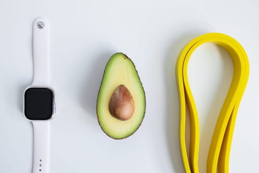 Minimalist arrangement with smartwatch, avocado, and yellow resistance bands on white background.