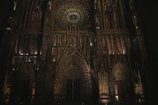 Symmetrical nighttime view of Strasbourg Cathedral Gothic facade, featuring the illuminated rose window and three ornate entrance portals.