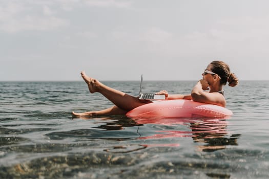 A woman is floating in a pink inflatable raft with a laptop in her hand. She is smiling and she is enjoying her time in the water