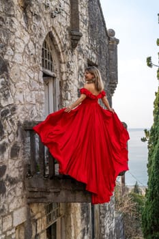 Woman in a flowing red gown stands on a stone castle balcony overlooking a sea view. The dress billows dramatically.