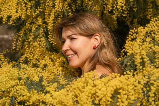 Smiling woman with shoulder-length blonde hair, wearing red earrings, surrounded by vibrant yellow mimosa flowers.