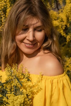 Blond woman in a yellow off-the-shoulder dress, gently smiling, holds a bouquet of mimosa flowers. Sunlight illuminates her face and the vibrant blossoms.