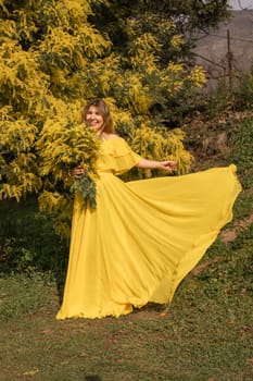 Woman in a flowing yellow gown stands near a mimosa bush, dress swirling, holding mimosa branches. Sunlight illuminates the scene.