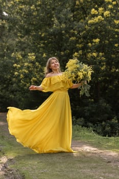 Woman in flowing yellow dress holds mimosa bouquet, standing in a sunlit path amidst blooming mimosa trees.
