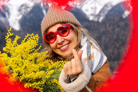 Smiling woman in heart-shaped sunglasses holds mimosa flowers, makes a heart sign with her fingers, mountainous backdrop.