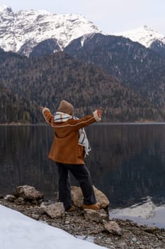 Woman in brown coat poses near a calm lake, snow-dusted rocks at her feet. Majestic snow-capped mountains form a dramatic backdrop.