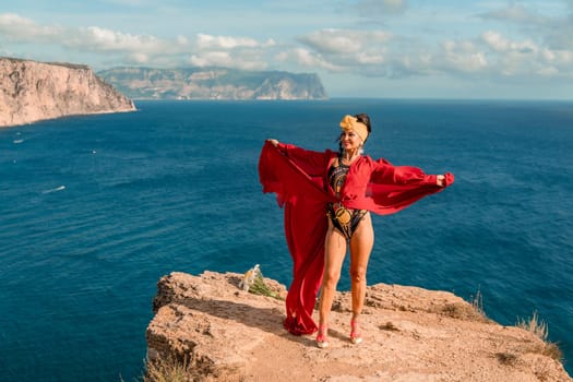 A woman in a red dress is standing on a rocky cliff overlooking the ocean. She is wearing a lei and she is dancing or celebrating. The scene is serene and peaceful, with the ocean as a backdrop