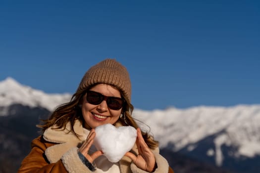 Smiling woman in sunglasses and winter attire holds a snow heart against a backdrop of snow-capped mountains under a clear blue sky.