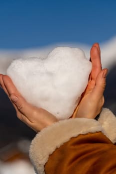 Two hands gently cradle a heart-shaped snowball against a vibrant blue sky and snowy mountain backdrop. Winter's delicate creation.