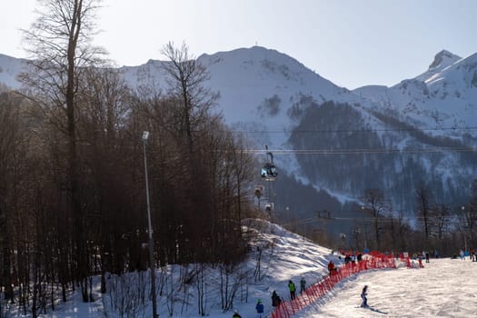 Sunny day at a snow-covered ski resort. People ski and snowboard down slopes, a chairlift ascends the mountainside, against a backdrop of snow-capped peaks and leafless trees.