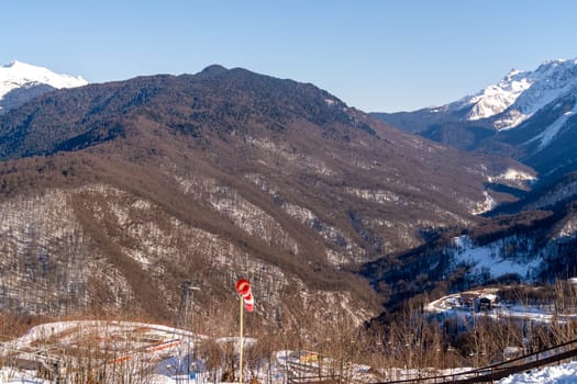 Panoramic view of snow-dusted mountains under a clear blue sky. A small flag stands on a pole in the foreground. Winter mountain landscape.